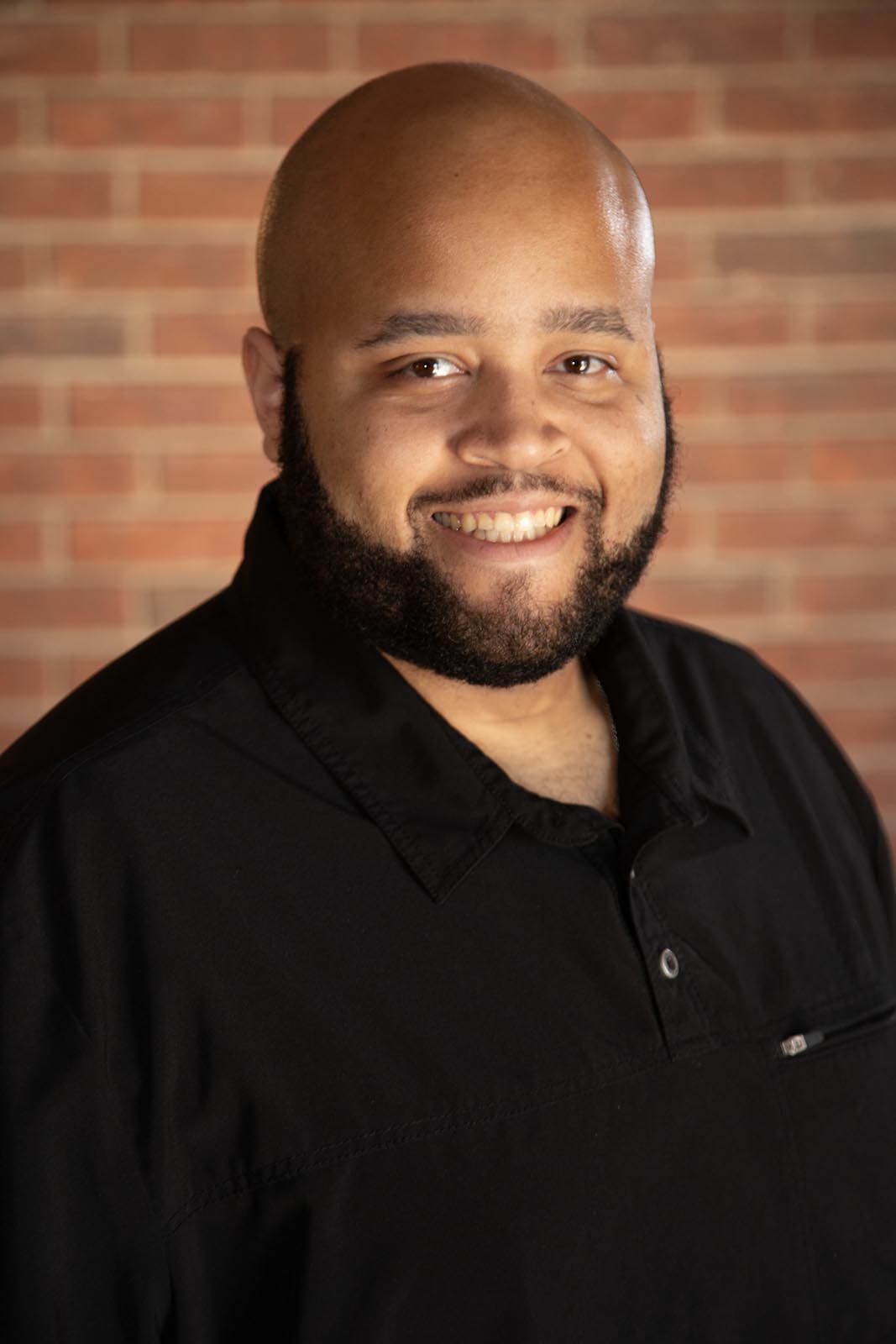 A person wearing a black collared shirt stands in front of a brick wall, with a buttoned closure and a small pocket on the left side. The textured brick background contrasts with the dark clothing, emphasizing a simple yet professional look.