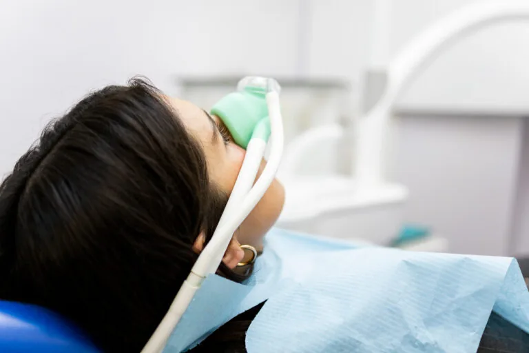 A person reclines in a dental chair wearing a blue bib, with a suction device or dental tool positioned in their mouth during a procedure. The background features dental equipment and a blurred clinical setting, reinforcing the professional environment of the treatment.